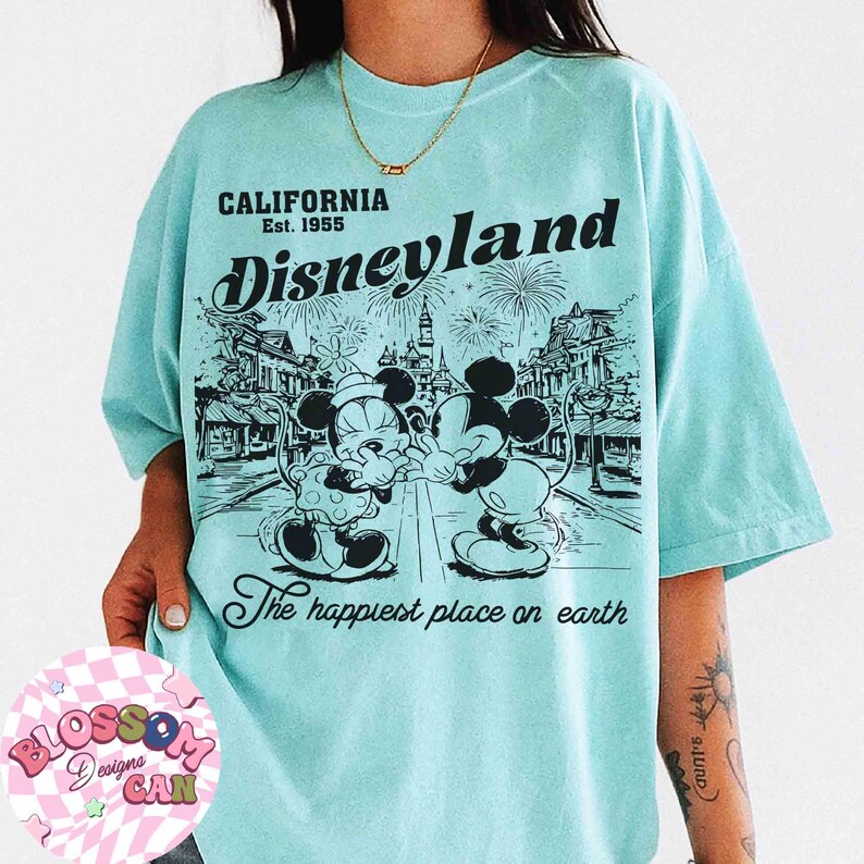 May include: A light blue t-shirt featuring a graphic of Mickey and Minnie Mouse in front of a Disneyland scene. The shirt includes the text "CALIFORNIA Est. 1955 Disneyland" and "The happiest place on earth."