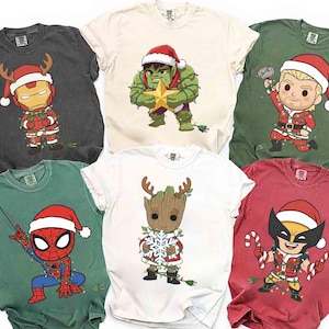 Könnte beinhalten: Sechs T-Shirts mit weihnachtlichen Superhelden-Designs. Die Shirts zeigen Charaktere wie Iron Man, Hulk, Thor, Spider-Man, Groot und Wolverine, alle mit Weihnachtsmützen und festlicher Kleidung. Jedes Shirt hat eine andere Farbe.