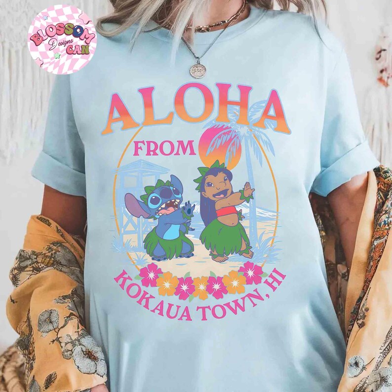 May include: Pale blue t-shirt with the words "Aloha From Kokaua Town, HI" in orange and pink. The shirt features a cartoon illustration of two characters in a tropical setting with palm trees and flowers. The shirt has rolled-up sleeves.