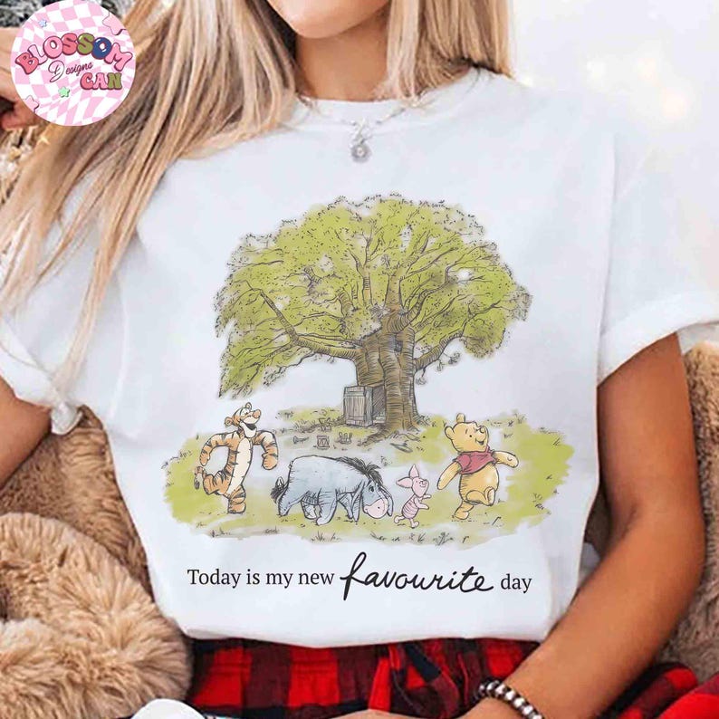 K&ouml;nnte beinhalten: Wei&szlig;es T-Shirt mit einer Illustration von Winnie Puuh, Tigger, Eeyore und Ferkel unter einem gro&szlig;en Baum. Der Text "Today is my new favourite day" ist in einer Schreibschrift geschrieben. Das Design verwendet weiche, ged&auml;mpfte Farben.