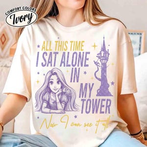 以下が含まれることがあります： アイボリー色のComfort Colors Tシャツ。長い髪の毛のキャラクター、塔、そして「ALL THIS TIME I SAT ALONE IN MY TOWER. Now I can see it all.」というテキストのグラフィックが特徴です。