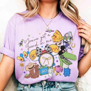 以下が含まれることがあります： 紫色のTシャツ。カラフルなグラフィックデザインで、「You've got a friend in me」の文字が入っています。デザインには、漫画のキャラクター、ロケット、カウボーイハット、緑色の恐竜が含まれています。
