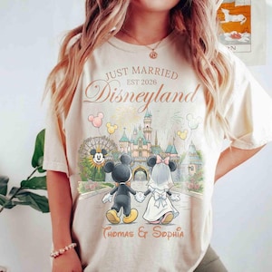 Może przedstawiać: Kremowa koszulka z motywem ślubu w Disneylandzie. Projekt przedstawia Myszki Miki i Minnie, zamek, fajerwerki oraz napis "Just Married Disneyland Est 2026" wraz z imionami. Koszulka ma luźny kr&oacute;j.