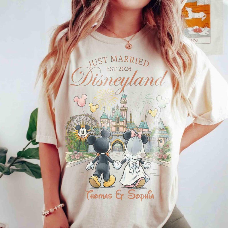 Może przedstawiać: Kremowa koszulka z motywem ślubu w Disneylandzie. Projekt przedstawia Myszki Miki i Minnie, zamek, fajerwerki oraz napis "Just Married Disneyland Est 2026" wraz z imionami. Koszulka ma luźny kr&oacute;j.