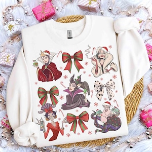 May include: White sweatshirt featuring cartoon illustrations of Disney villains, red and green bows, candy canes, and snowflakes. The design includes characters like the Evil Queen, Cruella de Vil, and Ursula, with festive holiday elements.