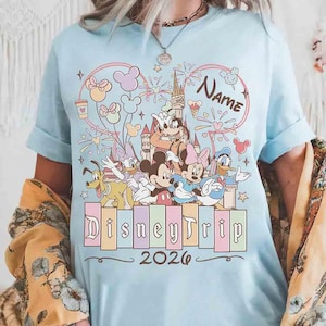 K&ouml;nnte beinhalten: Hellblaues T-Shirt mit einem farbenfrohen Disney Trip-Design. Das Design zeigt Micky Maus, Minnie Maus, Donald Duck und andere Charaktere sowie den Text "Disney Trip 2026" und das Wort "Name".
