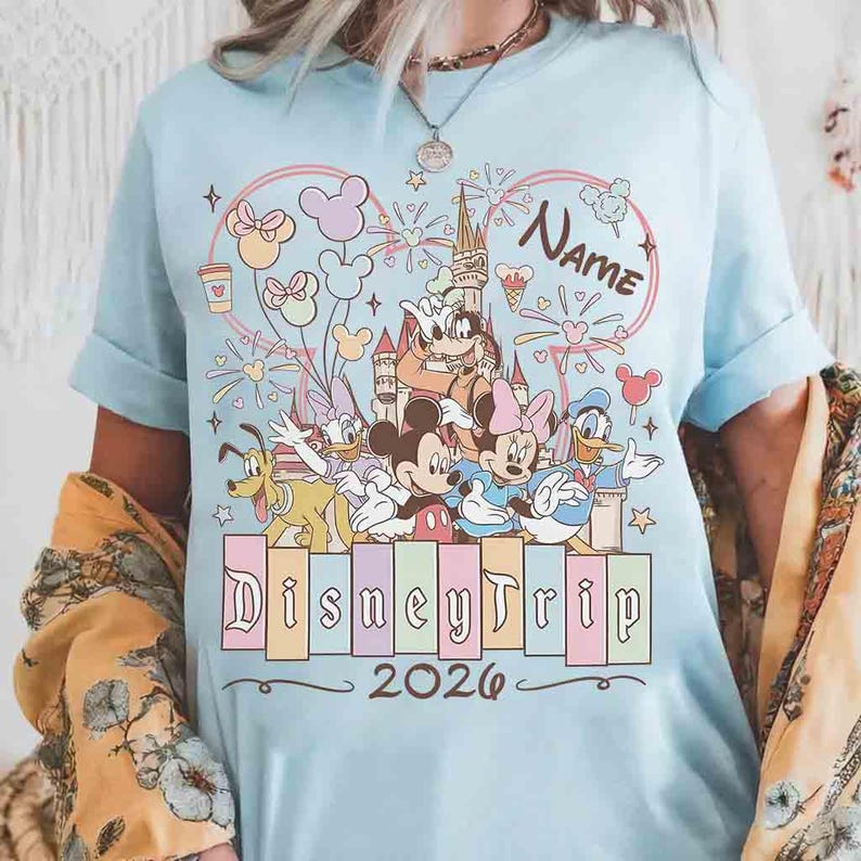 K&ouml;nnte beinhalten: Hellblaues T-Shirt mit einem farbenfrohen Disney Trip-Design. Das Design zeigt Micky Maus, Minnie Maus, Donald Duck und andere Charaktere sowie den Text "Disney Trip 2026" und das Wort "Name".