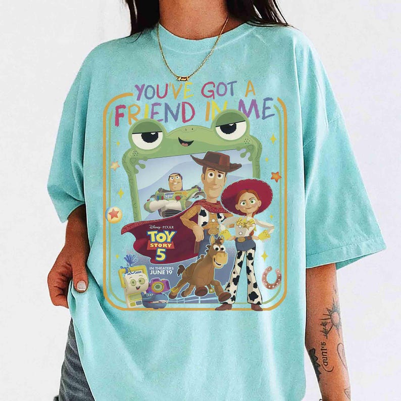 May include: A light blue t-shirt featuring a graphic print with the text "YOU'VE GOT A FRIEND IN ME" and "TOY STORY 5 IN THEATERS JUNE 19". The design includes characters Woody, Jessie, Buzz Lightyear, Bullseye, and others inside a green frog frame.