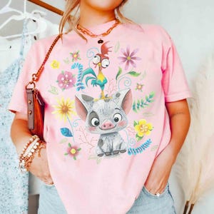 May include: A pale pink t-shirt with a cartoon illustration of a pig and a rooster, set amongst floral designs. The pig is grey and white, and the rooster is yellow, blue, and red. The shirt has a round neckline.