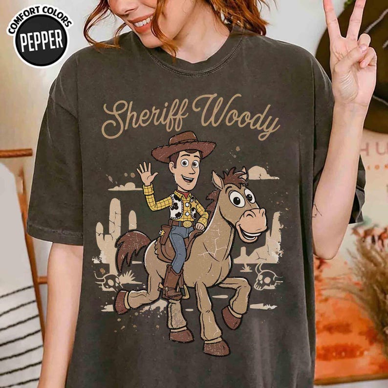 Op de afbeelding: Een antracietgrijs t-shirt met korte mouwen en een afbeelding van Sheriff Woody op het paard Bullseye. De tekst "Sheriff Woody" staat in een cursief lettertype boven de figuren. Op de achtergrond is een woestijnlandschap met cactussen te zien.