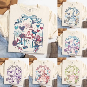 May include: Cream-colored t-shirts with a Disney theme. Each shirt features a graphic of Minnie Mouse and Daisy Duck in cheerleading outfits, a castle, fireworks, and the text "Walt Disney World". The design is framed by a blue ribbon.