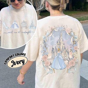 May include: Ivory-colored t-shirt featuring a Cinderella-themed design. The front displays a shoe, dress, carriage, and castle with the text "meet me at midnight." The back showcases Cinderella in front of a castle, surrounded by roses and bows.