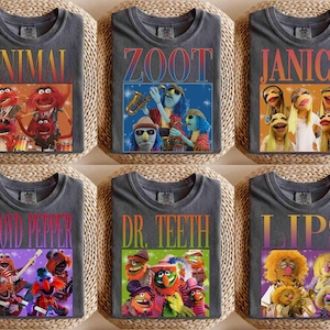 May include: Six charcoal gray t-shirts featuring Muppet characters and band members. Each shirt has a different name in a colorful font: Animal, Zoot, Janice, Floyd Pepper, Dr. Teeth, and Lips. Each shirt has a corresponding image of the character or band.