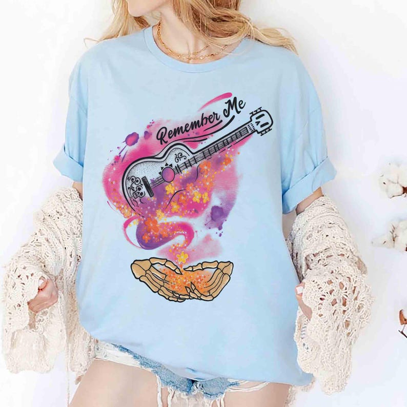 Puede incluir: Camiseta azul claro con un dise&ntilde;o gr&aacute;fico que presenta una guitarra, detalles florales y el texto "Remember Me". El dise&ntilde;o incorpora tonos rosas, morados y amarillos. La guitarra est&aacute; detallada con elementos en blanco y negro, con un estilo art&iacute;stico.