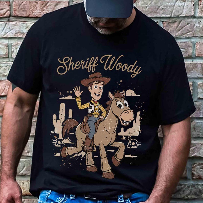 Op de afbeelding: Een zwart t-shirt met korte mouwen en een grafische print van het personage Sheriff Woody op een paard. Het ontwerp bevat een woestijnlandschap met cactussen en de tekst Sheriff Woody in een beige cursief lettertype erboven.