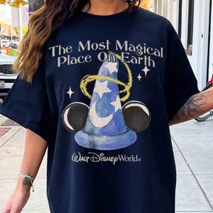 May include: Navy blue t-shirt with the text "The Most Magical Place On Earth" above a blue wizard hat with stars and a crescent moon. The "Walt Disney World" logo is below the hat, with black Mickey Mouse ears on either side.