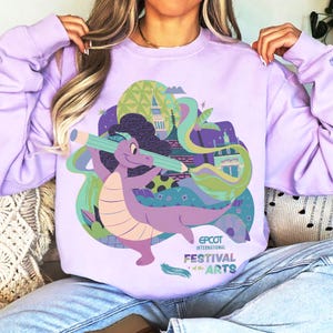 Puede incluir: Sudadera lila con un dibujo de un dragón morado sosteniendo un lápiz verde, sobre un fondo de edificios y formas coloridas. El texto "EPCOT INTERNATIONAL FESTIVAL of the ARTS" está impreso debajo.