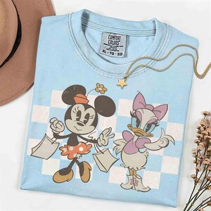 May include: Light blue Comfort Colors t-shirt featuring Minnie Mouse and Daisy Duck with shopping bags. The shirt has a checkered background and a gold star necklace. The tag reads XL-TG-EG.