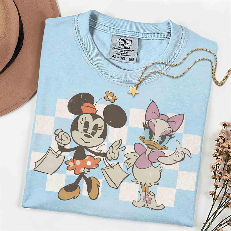 May include: Light blue Comfort Colors t-shirt featuring Minnie Mouse and Daisy Duck with shopping bags. The shirt has a checkered background and a gold star necklace. The tag reads XL-TG-EG.