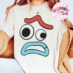 May include: A white t-shirt featuring a cartoon face with a red curved line above two eyes, a large black circle and a smaller black circle, and a blue open mouth. The shirt has the words "Blossom Designs CAN" in a pink circle.