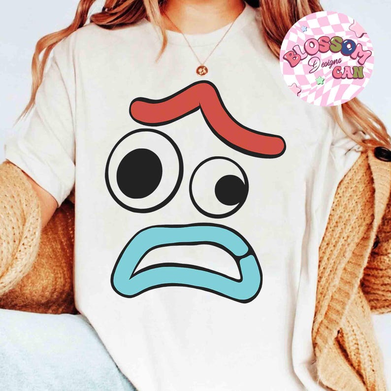 May include: A white t-shirt featuring a cartoon face with a red curved line above two eyes, a large black circle and a smaller black circle, and a blue open mouth. The shirt has the words "Blossom Designs CAN" in a pink circle.