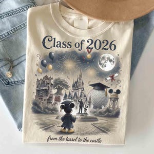 May include: Cream-colored t-shirt featuring a graduation-themed illustration with Mickey Mouse in a cap and gown, fireworks, and the text "Class of 2026" and "from the tassel to the castle".