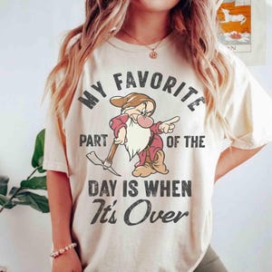 May include: A cream-coloured t-shirt with a cartoon dwarf holding a pickaxe. The text reads "MY FAVORITE PART OF THE DAY IS WHEN It's OVER" in black. The dwarf is dressed in red with a brown hat.