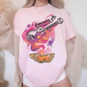Puede incluir: Camiseta rosa claro con un dise&ntilde;o de guitarra y la frase "Remember Me". La guitarra se encuentra sobre un fondo de acuarela colorido con acentos naranjas y morados. El dise&ntilde;o incluye un par de manos esquel&eacute;ticas.