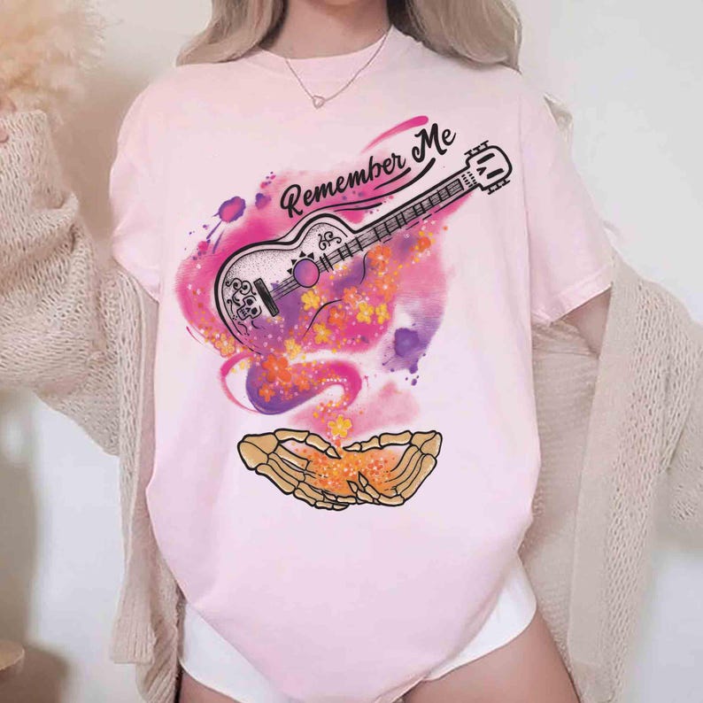 Puede incluir: Camiseta rosa claro con un dise&ntilde;o de guitarra y la frase "Remember Me". La guitarra se encuentra sobre un fondo de acuarela colorido con acentos naranjas y morados. El dise&ntilde;o incluye un par de manos esquel&eacute;ticas.