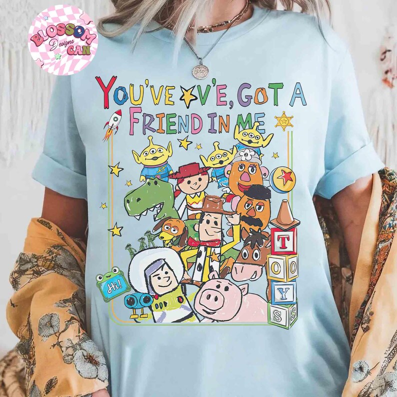 May include: Light blue t-shirt featuring a colorful graphic of characters and the text "YOU'VE GOT A FRIEND IN ME". The design includes a rocket, stars, and various cartoon figures. The shirt is a casual wear item.