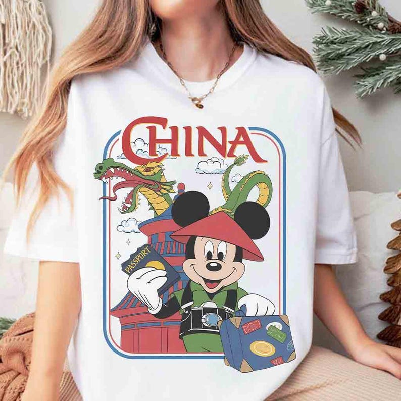 Pode incluir: Camiseta branca com um desenho do Mickey Mouse em traje chin&ecirc;s, segurando um passaporte e uma mala. O design inclui um drag&atilde;o, uma pagoda e a palavra "CHINA" em letras vermelhas.