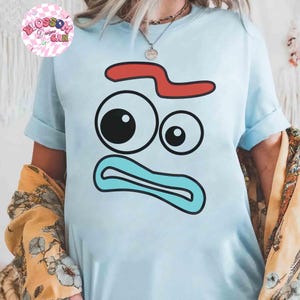 May include: Light blue t-shirt featuring a cartoon face design with a red curved shape, two eyes, and a blue mouth. The shirt has rolled-up sleeves and the text "Blossom Designs Can" in a pink circle.