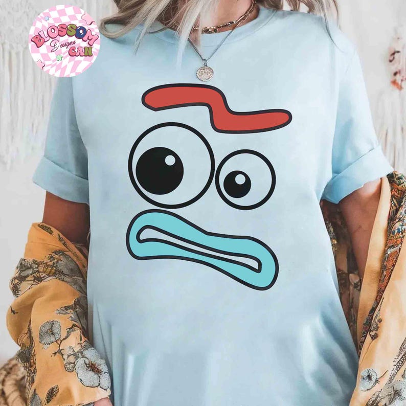 May include: Light blue t-shirt featuring a cartoon face design with a red curved shape, two eyes, and a blue mouth. The shirt has rolled-up sleeves and the text "Blossom Designs Can" in a pink circle.