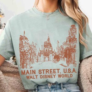 Peut inclure: T-shirt vert clair avec une illustration marron de Main Street, U.S.A. &agrave; Walt Disney World. Le motif comprend des b&acirc;timents, un ch&acirc;teau et le texte "MAIN STREET, U.S.A. WALT DISNEY WORLD" en gras.