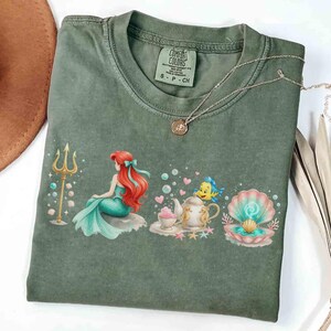 May include: An olive green Comfort Colors t-shirt featuring a graphic of a mermaid with red hair, a golden trident, and a seashell. A silver necklace with a pendant is draped across the shirt. The shirt is size small.