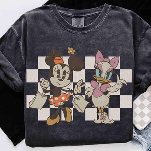 May include: Dark gray t-shirt featuring Minnie Mouse and Daisy Duck with shopping bags, set against a black and white checkered background. Minnie Mouse wears a polka dot skirt and Daisy Duck wears a pink bow and dress. The shirt has a vintage, distressed look.