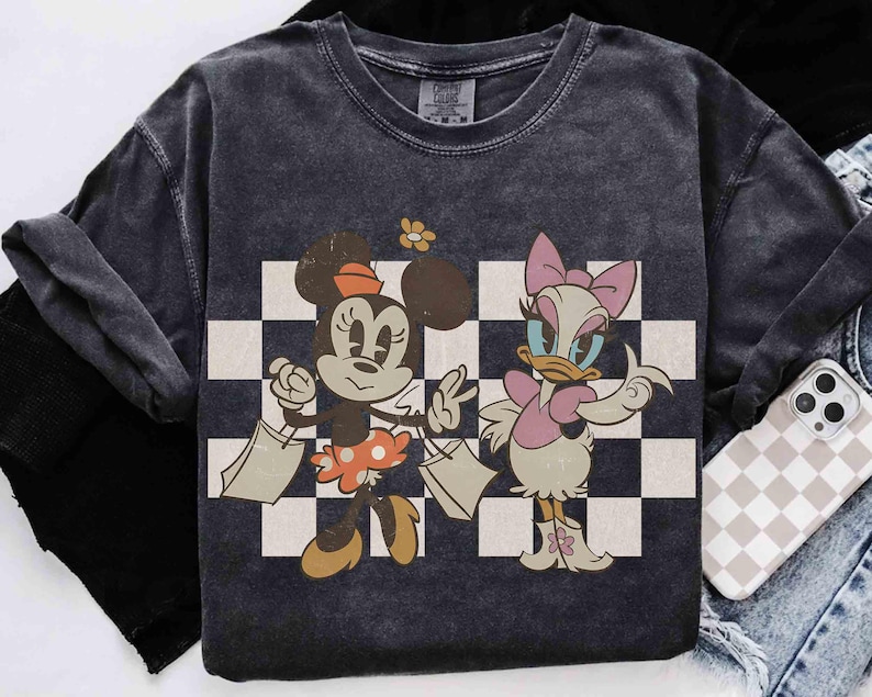 May include: Dark gray t-shirt featuring Minnie Mouse and Daisy Duck with shopping bags, set against a black and white checkered background. Minnie Mouse wears a polka dot skirt and Daisy Duck wears a pink bow and dress. The shirt has a vintage, distressed look.