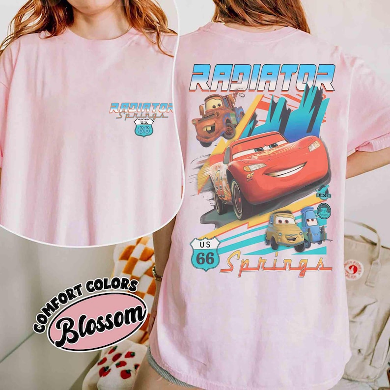 Puede incluir: Una camiseta rosa claro con un peque&ntilde;o logotipo de Radiator Springs U.S. 66 en el pecho. La parte posterior muestra un gr&aacute;fico grande de Lightning McQueen, Mater y otros personajes con el texto Radiator Springs y U.S. 66.