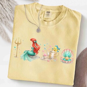 May include: Yellow t-shirt featuring a mermaid with red hair and a teal tail, sitting on a rock. A silver necklace with a pendant rests on the shirt. The shirt also has a teapot, teacup, and seashell graphic.