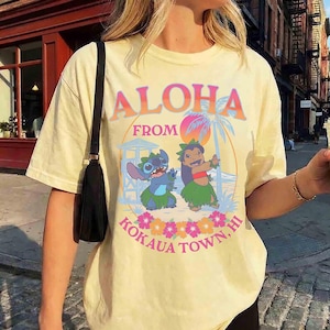 May include: Pale yellow t-shirt featuring the text "ALOHA FROM KOKAUA TOWN, HI" in a colourful design. The graphic includes cartoon characters, a palm tree, and flowers. The shirt has short sleeves.