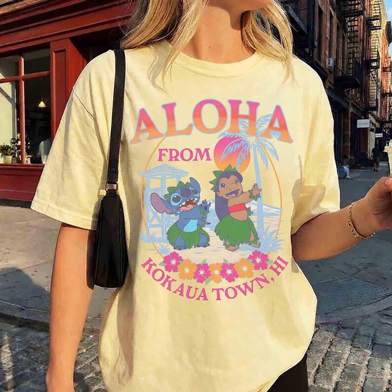 May include: Pale yellow t-shirt featuring the text "ALOHA FROM KOKAUA TOWN, HI" in a colourful design. The graphic includes cartoon characters, a palm tree, and flowers. The shirt has short sleeves.