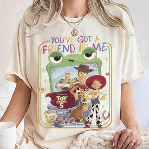 May include: A cream-colored t-shirt featuring a graphic print with the text "YOU'VE GOT A FRIEND IN ME" and "TOY STORY 5". The design includes characters Woody, Buzz Lightyear, Jessie, Bullseye, and Forky framed by a green frog-shaped border.
