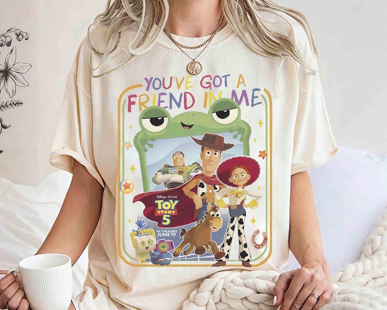 May include: A cream-colored t-shirt featuring a graphic print with the text "YOU'VE GOT A FRIEND IN ME" and "TOY STORY 5". The design includes characters Woody, Buzz Lightyear, Jessie, Bullseye, and Forky framed by a green frog-shaped border.
