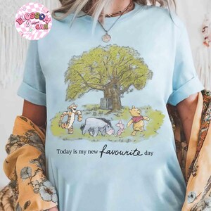K&ouml;nnte beinhalten: Hellblaues T-Shirt mit einer Grafik von Winnie Puuh und seinen Freunden unter einem Baum. Der Text "Today is my new favourite day" ist unter dem Bild aufgedruckt. Das Shirt hat hochgekrempelte &Auml;rmel.