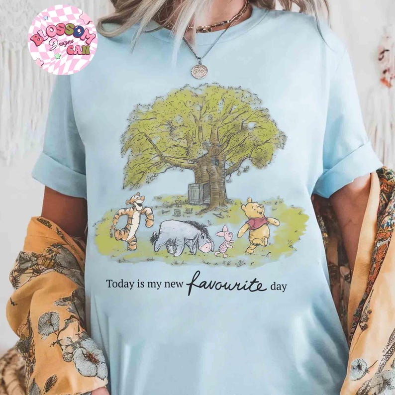 K&ouml;nnte beinhalten: Hellblaues T-Shirt mit einer Grafik von Winnie Puuh und seinen Freunden unter einem Baum. Der Text "Today is my new favourite day" ist unter dem Bild aufgedruckt. Das Shirt hat hochgekrempelte &Auml;rmel.