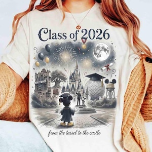 May include: Cream-colored t-shirt featuring a graduation-themed graphic. The design includes the text "Class of 2026" above a detailed illustration of a castle, fireworks, and Mickey Mouse in a graduation gown. The phrase "from the tassel to the castle" is printed at the bottom.