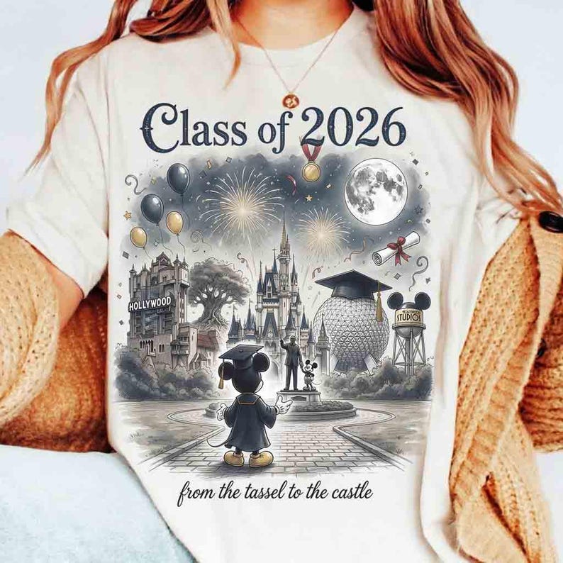 May include: Cream-colored t-shirt featuring a graduation-themed graphic. The design includes the text "Class of 2026" above a detailed illustration of a castle, fireworks, and Mickey Mouse in a graduation gown. The phrase "from the tassel to the castle" is printed at the bottom.