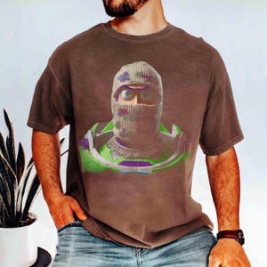 May include: A brown short-sleeve t-shirt featuring a graphic print of a character wearing a camouflage-patterned knit balaclava, a silver chain necklace, and a green and purple space-themed suit with a high collar and shoulder armor pieces.