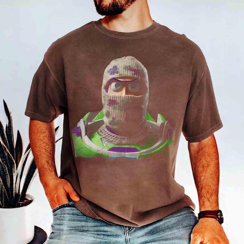 May include: A brown short-sleeve t-shirt featuring a graphic print of a character wearing a camouflage-patterned knit balaclava, a silver chain necklace, and a green and purple space-themed suit with a high collar and shoulder armor pieces.
