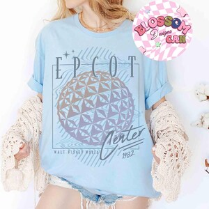 May include: Light blue t-shirt featuring a graphic of the EPCOT Center sphere with the text "EPCOT Center Walt Disney World 1982". The design is in shades of gray and purple. The shirt has rolled-up sleeves.